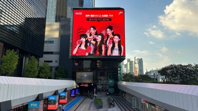 Billboard featuring NewJeans music group promoting Coca-Cola in an urban cityscape.