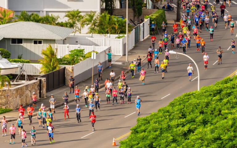 Runners participating in a marathon on a sunny day, with residential homes and green trees in the background.