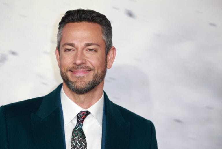 Zachary Levi wearing a teal blazer and patterned tie, smiling against a light, blurred background.