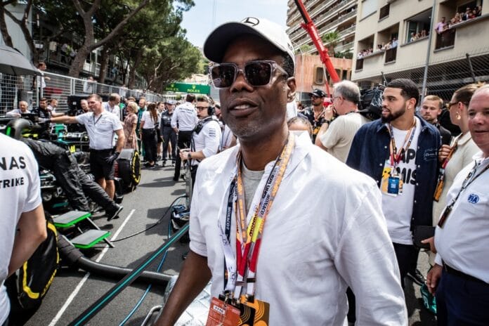 Chris Rock at the Monaco Grand Prix, wearing sunglasses and a white shirt, surrounded by a crowd and Formula 1 cars in the background.