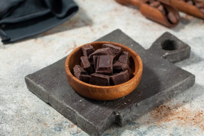 A wooden bowl filled with chunks of dark chocolate on a rustic slate cutting board, with a blurred background of cocoa scoops.