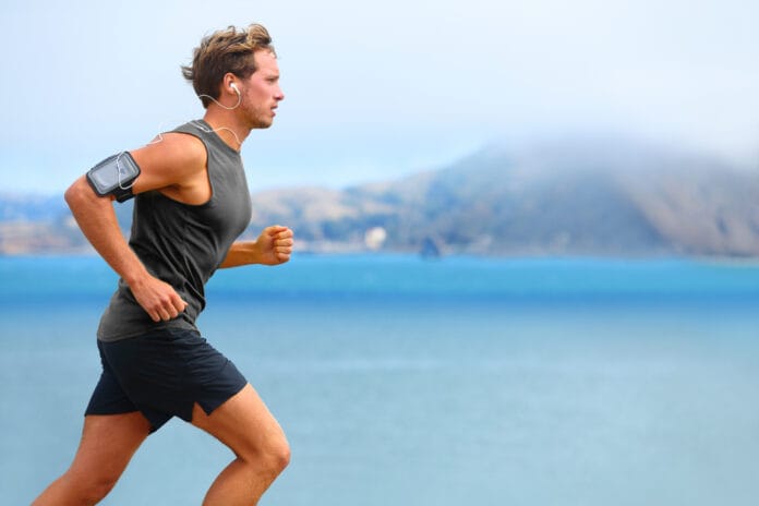 Man running outdoors near water with mountains in the background, wearing a black tank top, shorts, and an armband with a phone.