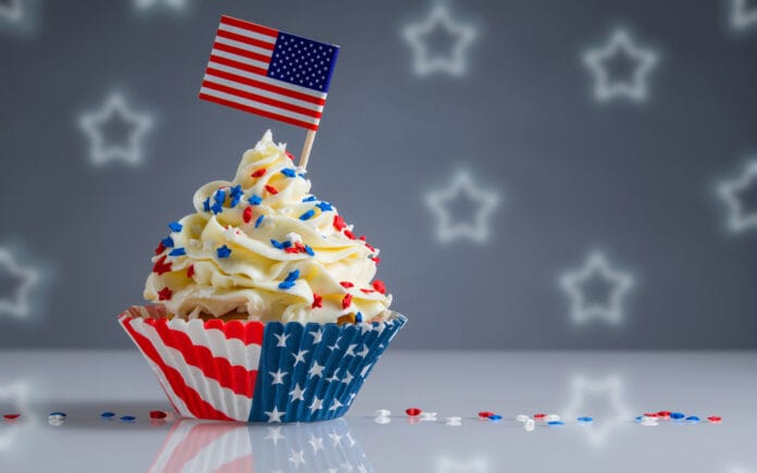 A patriotic cupcake decorated with red, white, and blue sprinkles, topped with an American flag, in a wrapper featuring the U.S. flag design. The background has a blurred star pattern.