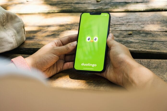 A person holding a smartphone with the Duolingo app open on the screen, featuring the green owl mascot, sitting outdoors on a wooden surface.