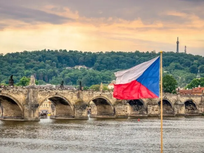 Charles Bridge in Prague at sunset, with the Czech flag fluttering in the breeze, river Vltava below, and Petřín hill in the background, inspiring a taste of Czech dishes.