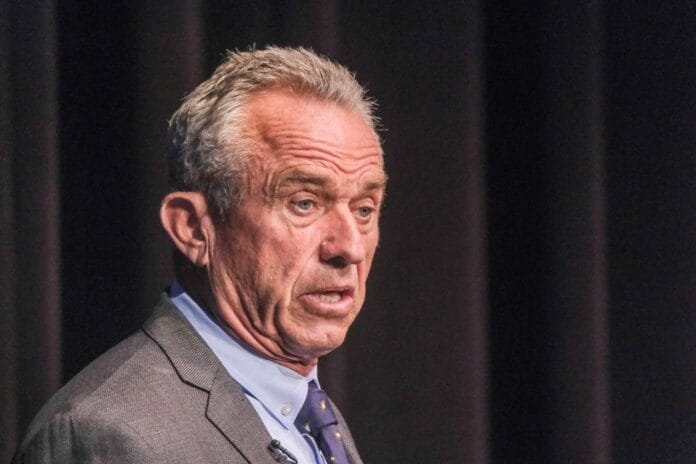 Robert F. Kennedy Jr. delivering a speech on stage, wearing a suit and tie, with a serious expression.