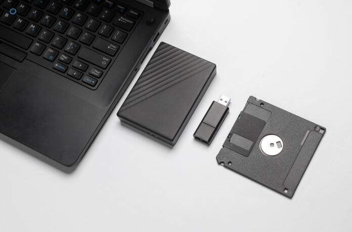 A black laptop with various storage devices, including an external hard drive, USB flash drive, and a floppy disk, representing the evolution of data storage.