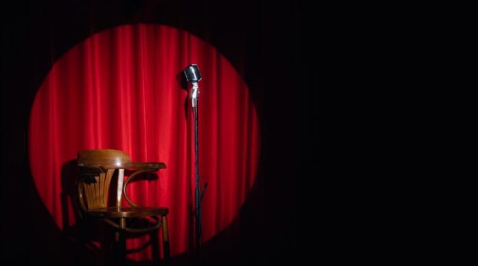 Empty comedy stage with a vintage microphone, wooden chair, and red curtains under a spotlight.