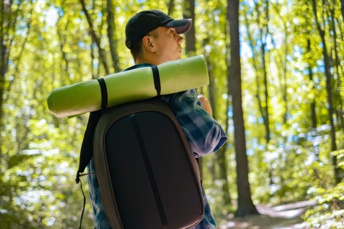 A man hiking through a sunlit forest with a backpack and a rolled-up mat, illustrating the growing fitness trend of rucking.