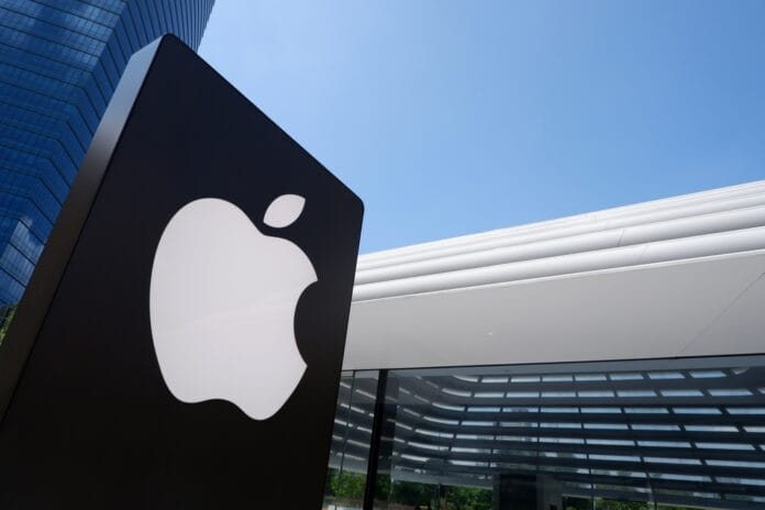 Apple logo on a black sign in front of a modern glass and metal building with a blue sky in the background.