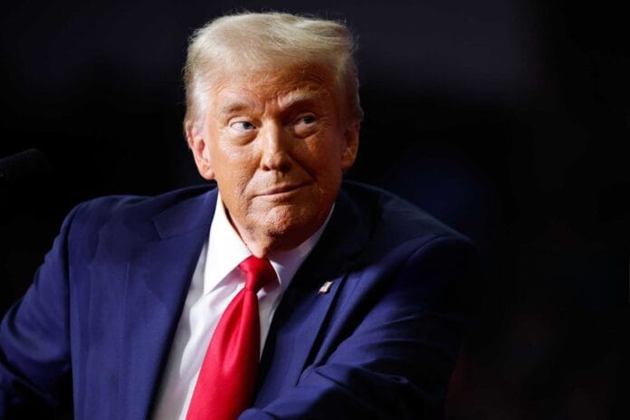 Donald Trump speaking at an event, wearing a blue suit and red tie with a focused look.